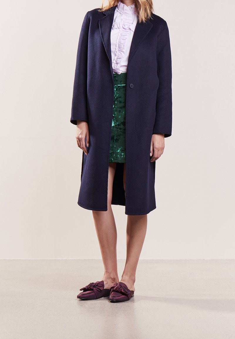 Manteau en laine marine à manches longues et à l'avant ouvert, porté sur une jupe verte à sequins, associé à des chaussures violettes à enfiler avec un détail en nœud.