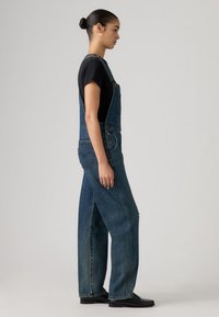 Γυναίκα με μαλλιά σε κότσο φοράει μαύρο μπλουζάκι, σκούρα μπλε τζιν σαλόπες και μαύρα μοκασίνια, στέκεται πλευρικά μπροστά σε απλό φόντο.