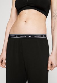 Melni bikses ar mīkstu tekstūru, ar melnu jostasvietu, kurā ir balts "LACOSTE" logotips un krokodila akcents. Šaura piegriezuma dizains.