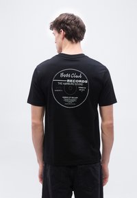 Άνδρας που φοράει μαύρο μπλουζάκι με μεγάλο λευκό γραφικό ετικέτας δίσκου και κείμενο "Boss Club Records The Hamburg Sound" στην πλάτη, στέκεται μπροστά σε λευκό φόντο.