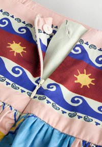 Gros plan sur un tissu coloré avec des motifs de soleil et de vagues, comprenant une fermeture éclair partiellement ouverte et un bouton blanc sur un tissu rose.