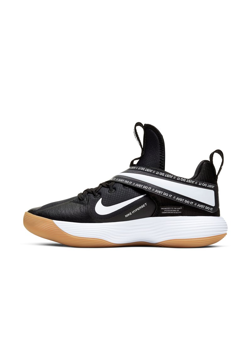 Scarpa sportiva nera con tomaia testurizzata, logo Nike bianco e suola in gomma. Presenta un cinturino con branding ben visibile con il testo "Just Do It".