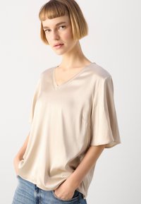 Beige satintopp med V-hals og korte ermer, med en løs passform og sidelomme, kombinert med lyseblå jeans.