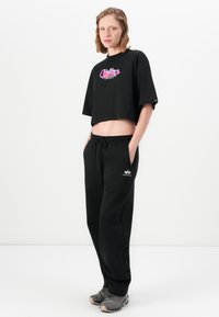Kvinde står med hænderne i lommerne iført sort cropped graphic T-shirt, sorte drawstring sweatpants og grå sportsko imod hvid baggrund.