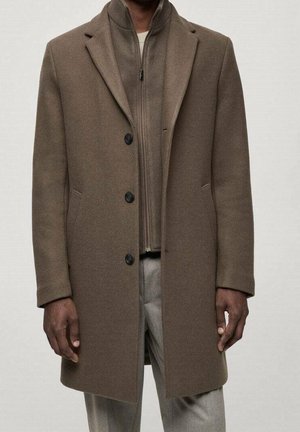 Mężczyzna ubrany w brązowy, zapinany na guziki płaszcz na kurtkę zapinaną na zamek oraz beżowe spodnie, stojący na tle gładkiego, jasnego tła.