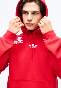 Noor punane kapuutsiga jope, millel on valge Adidas logo ja graafika, kohandab kapuutsi mõlema käega, vaadates pooleldi küljele.