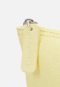 Gul dragkedjefodral gjort av texturerat material, med en präglad tag från Lacoste och silverfärgad hårdvara för stängning.