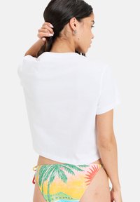 White cropped t-shirt with short sleeves, paired with colorful bikini featuring palm trees and sun patterns in yellow, pink, and green.