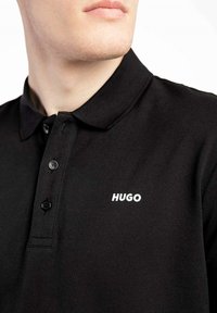 Chemise polo noire avec col, deux boutons, tissu texturé et logo "HUGO" blanc sur le côté gauche.