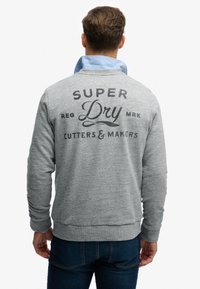 Grijze sweatshirt met een gestructureerde afwerking, voorzien van een blauwe kraag, bedrukt logo op de achterkant, en een geribbelde zoom.