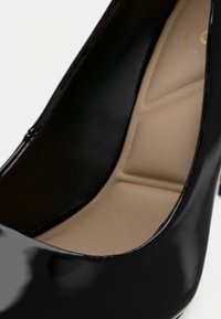Escarpin en cuir verni noir avec une finition lisse et brillante ; l'intérieur présente une semelle rembourrée beige avec un motif géométrique.
