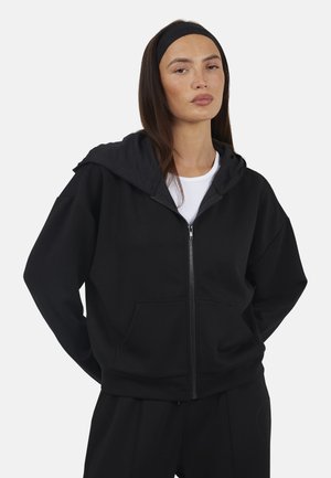 Woman with long dark hair and black headband wearing a black zip-up hoodie over a white shirt, standing with hands behind her back.