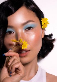 Una persona con pelle di tonalità media e capelli scuri e mossi, che tiene fiori gialli, indossa ombretto blu e una maglietta bianca su uno sfondo chiaro.
