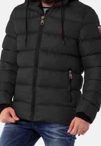 Giacca imbottita nera con chiusura a zip, cappuccio regolabile e due tasche laterali. Presenta polsini a costine e una toppa con il logo sulla manica.