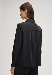 Zwarte lange mouwen shirt gemaakt van gladde stof. Heeft een kraag, knoopmanchetten en een geplooide achterkant voor extra val.