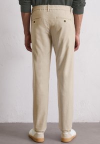 Homme portant un pantalon beige avec des poches arrières à boutons et des baskets blanches, debout contre un mur texturé blanc.