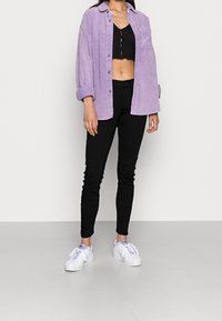 Camisa de pana lila de corte oversized, jeans ajustados negros y zapatillas blancas. La camisa tiene un diseño de botonadura y un bolsillo en el pecho.