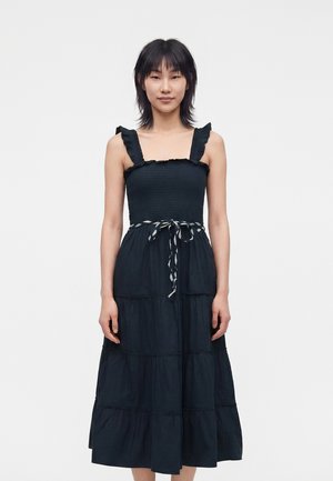 Femme debout de face, portant une robe bleu marine sans manches à volants avec bretelles à froufrous et une ceinture nouée dans un studio sobre.