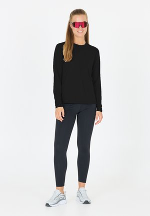 Black long-sleeve top, dark leggings, and silver athletic shoes. Model wearing large pink sunglasses, light hair styled in loose waves.