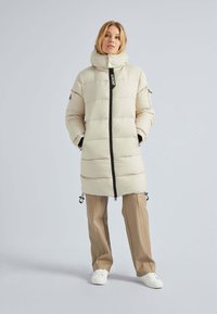 Manteau matelassé beige avec un col montant, une fermeture éclair noire et des ourlets à cordon de serrage. Porté par-dessus un pantalon beige et des baskets blanches. Texture lisse.