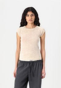 Beige short-sleeve top, lightweight fabric with a faded texture. Round neckline and fitted design, paired with gray sweatpants.
