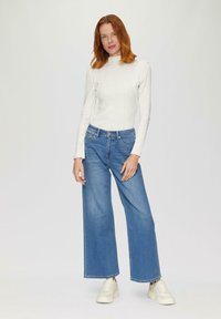 Haut à manches longues blanc avec un col montant, associé à un jean en denim bleu à jambes larges. Le modèle porte des baskets blanches. Fond uni.