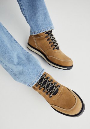 Bottes à lacets en daim beige avec des lacets noirs et blancs, portées avec un jean en denim bleu clair, sur fond blanc.