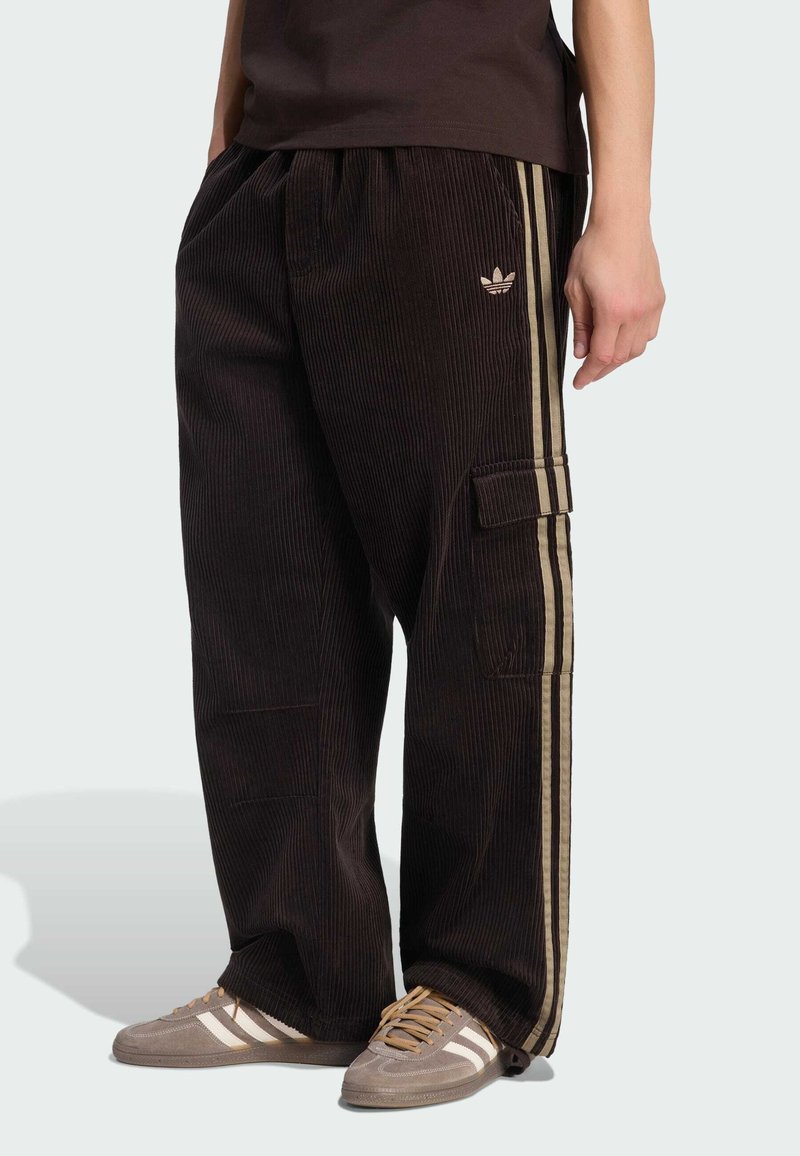 Personne portant un pantalon en velours côtelé noir avec des bandes latérales beiges et le logo Adidas, associé à des baskets Adidas grises avec des lacets beiges.