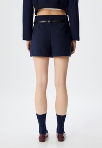 Lyhytlahkeiset tummansiniset housut, joissa on sileä pinta, musta vyötärönauha ja laskokset, yhdistetty tummiin sukkiin ja ruskeisiin krokotiilikuosisempiin kenkiin.