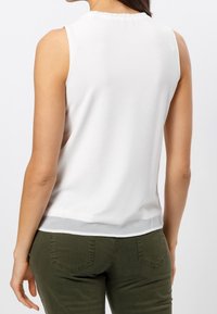Frau in ärmellosem weißen Top und olivgrüner Hose, von hinten vor einem schlichten hellen Hintergrund gesehen.