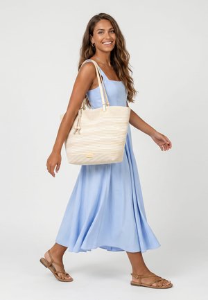 Donna sorridente in abito azzurro senza maniche che cammina, con grande borsa beige, indossa sandali bassi marroni su sfondo bianco.