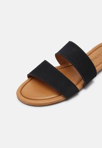 Sandalo piatto con suola color cuoio e due ampie cinghie in suede nero che attraversano il piede, esposto su uno sfondo bianco.