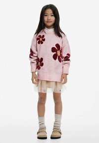 Svetloružový pletený sveter s červenými kvetmi, navrchu priehľadnej bielej sukne. Modelka má na nohách béžové topánky a hnedé pletené ponožky.