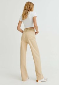 Femme avec de longs cheveux ondulés portant un pantalon large beige, un crop top blanc et des baskets blanches, se tenant contre un fond clair.