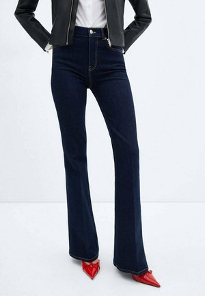 Personne portant un jean évasé taille haute bleu foncé, une veste en cuir noire, une chemise blanche et des talons rouges à bout pointu, debout les mains derrière le dos.