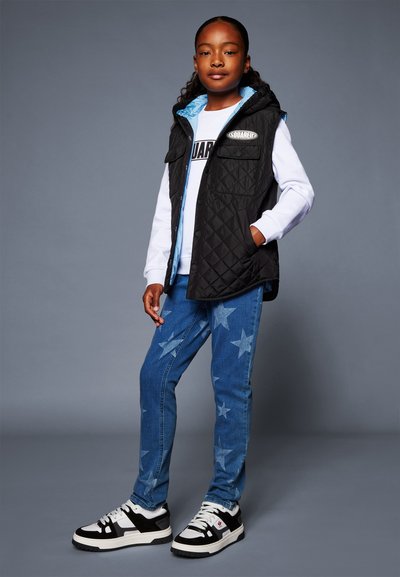 Gilet imbottito nero con fodera blu, camicia a maniche lunghe con grafica bianca, jeans blu con motivo a stelle e sneakers bianco e nere.