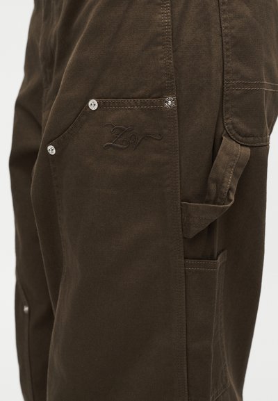 Καφέ cargo παντελόνι λεπτομέρεια με ασημί πριτσίνια, πλευρική τσέπη, θηλιά για εργαλεία και διακριτικό κεντητό λογότυπο στο ύφασμα.