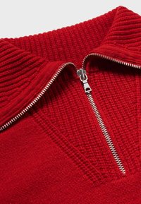 Pull rouge en tricot avec un col montant, doté d'une fermeture éclair en métal. La texture est côtelée près du col, tandis que le corps est lisse.
