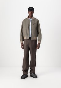 Jachetă bomber verde măslin cu un buzunar cu fermoar, asortată cu pantaloni maro. O caschetă neagră și adidași gri-negru completează ținuta.