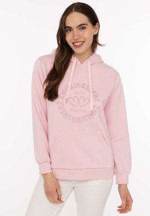 Frau mit langen braunen Haaren, die einen hellrosa Hoodie mit Herzgrafiken und Text trägt, kombiniert mit weißen Hosen, lächelt in die Kamera.