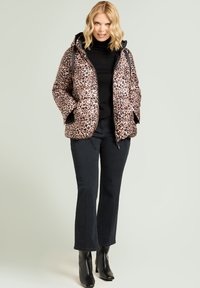 Chaqueta acolchada con estampado de leopardo, capucha, cremallera frontal y dos bolsillos laterales. Llevada sobre un suéter de cuello alto negro y combinada con jeans negros acampanados.