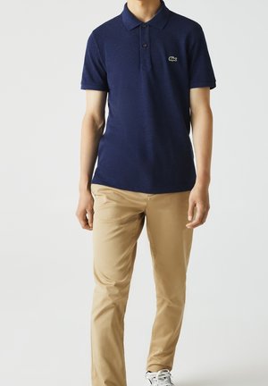 Mann trägt ein dunkelblaues Polohemd mit Logo, beige Hosen und weiße Sneakers und steht vor einem schlichten Hintergrund.