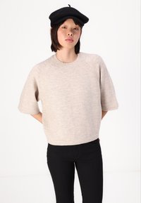 Un pull beige clair à manches courtes avec un col rond et des bords côtelés, associé à un pantalon noir. Le tissu semble doux et texturé.