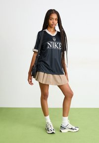 Svart Nike sportsjersey med hvite detaljer og logo, kombinert med en beige plissert skjørt og hvite crew-sokker. Sko med flerfargede detaljer.
