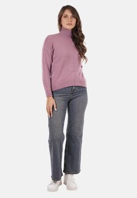 Donna con lunghi capelli ricci, che indossa un maglione viola a collo alto, jeans blu e sneakers bianche, in piedi davanti a uno sfondo semplice.