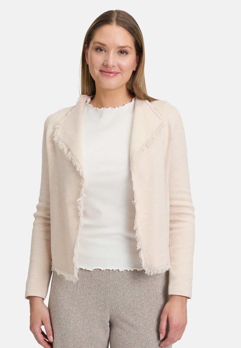 Cardigan rose clair avec des franges le long de l'ouverture et des manches, fabriqué en tissu doux. Porté sur un haut blanc et un pantalon à motifs.