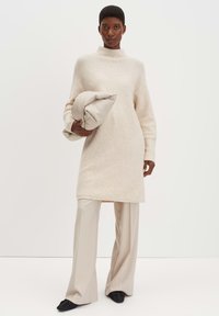 Strickkleid in Hellbeige mit hohem Kragen, langen Ärmeln und seitlichen Schlitzen, kombiniert mit beigen weit geschnittenen Hosen und schwarzen Schuhen.