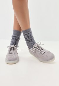 Tenisi gri din suede, cu bot rotund și șireturi, asortați cu șosete texturate gri cu dungă, așezate casual pe un fundal deschis.