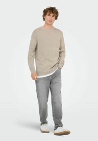 Beige Strickpullover mit strukturiertem Muster, Rundhalsausschnitt, kombiniert mit grauen Jeans und weißen Sneakers. Lässiger, bequemer Schnitt.
