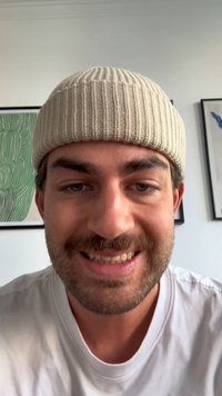 Un primer plano de una persona que lleva un gorro de canalé beige claro y una camiseta blanca, sonriendo, con un fondo de pared neutro y arte enmarcado.